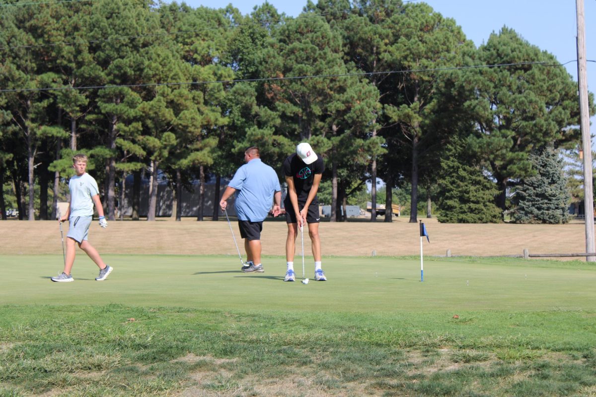 Junior, Braylen Turner, lines up putt before practice. While, Sophomore, Caleb Hagarty watches and Coach Travis Kuhn walks in the background.